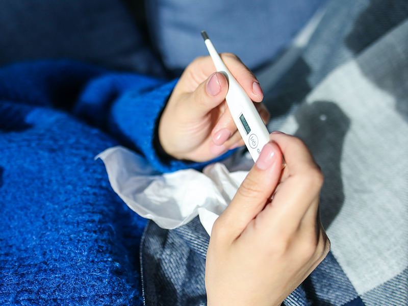 Person in a blue sweater holding a thermometer and a tissue.