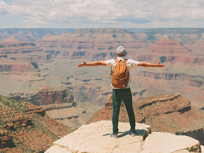 A person wearing an orange backpack stands at the rim of the Grand Canyon with arms spread.