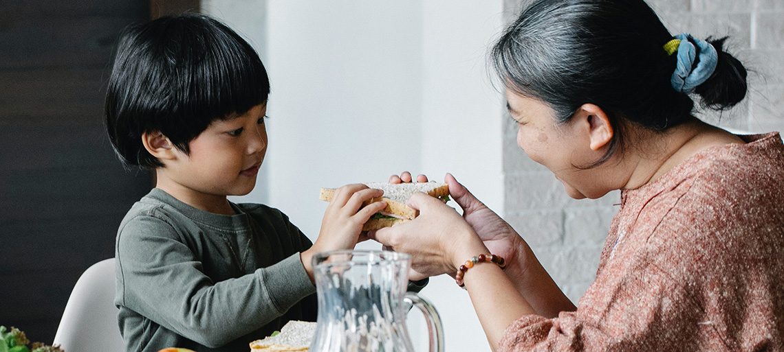 An older woman offers a sandwich to a young child at a table. She has dark hair and is smiling.