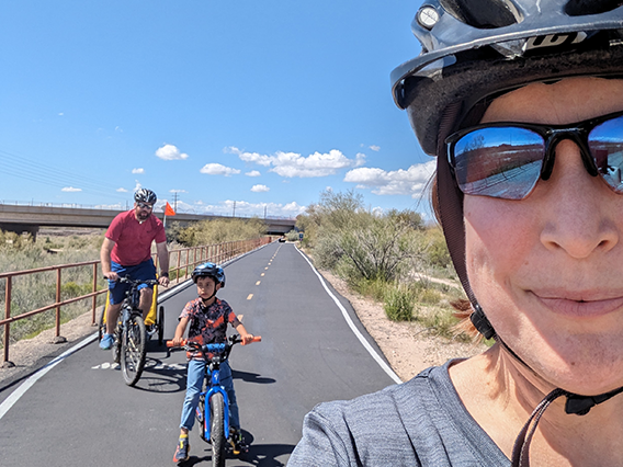 Foreground of a woman wearing a bike helmet taking a selfie with a man and boy each on a bike