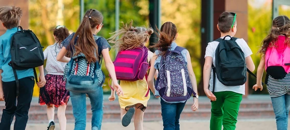 Group of elementary school children with colorful backpacks running together toward school building on a sunny day