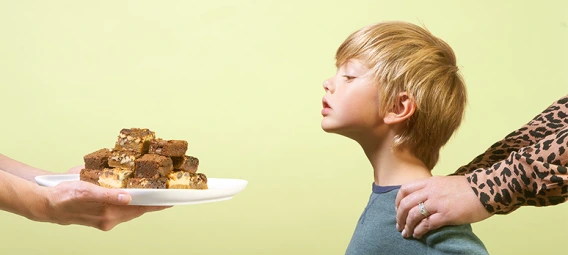 Parent's hand offering plate of cookies to young child who appears hesitant, representing food allergy awareness and dietary restrictions.