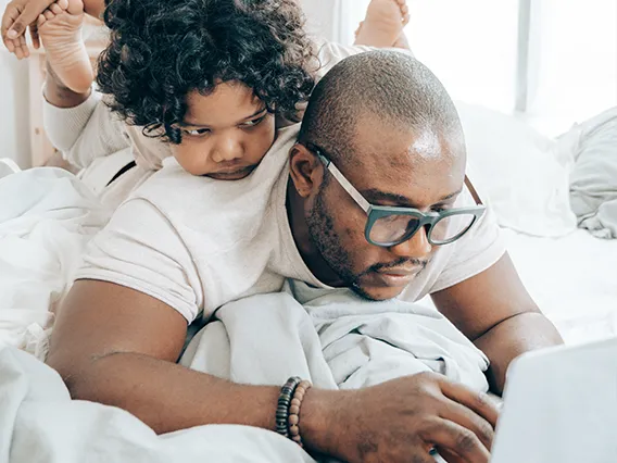 a father laying on a bed working on her computer with his daughter on this back