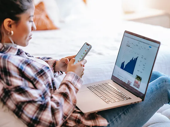 A woman with a laptop on her lap looking at financials while holding her cellphone