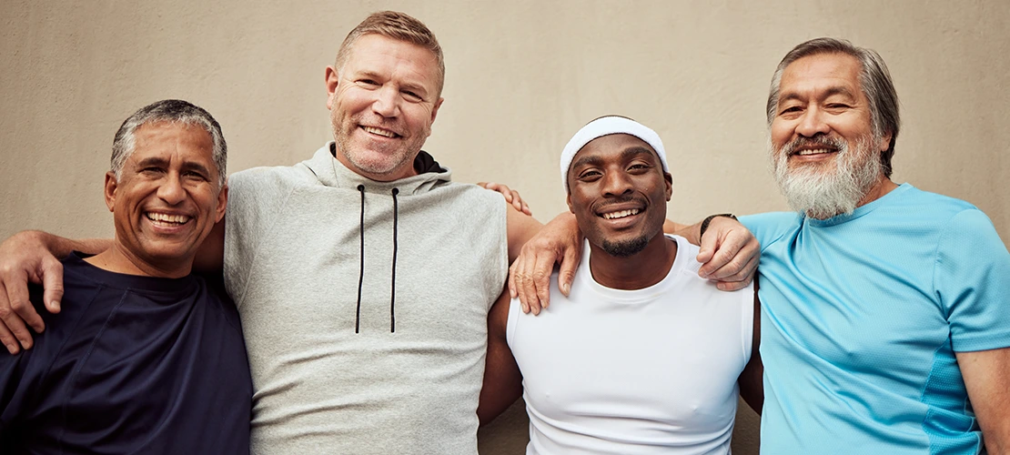 Four smiling men in athletic wear stand close together with their arms around each other, posing happily in front of a plain wall.
