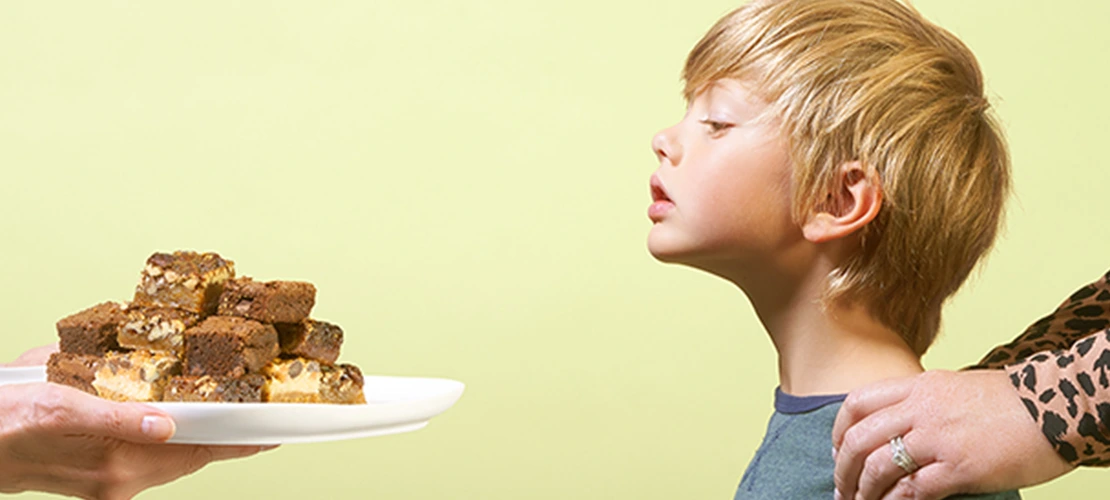 Parent's hand offering plate of cookies to young child who appears hesitant, representing food allergy awareness and dietary restrictions.