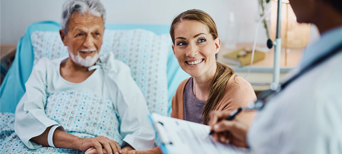 A smiling elderly man in a hospital bed holds hands with a young woman as they talk to a doctor holding a clipboard.