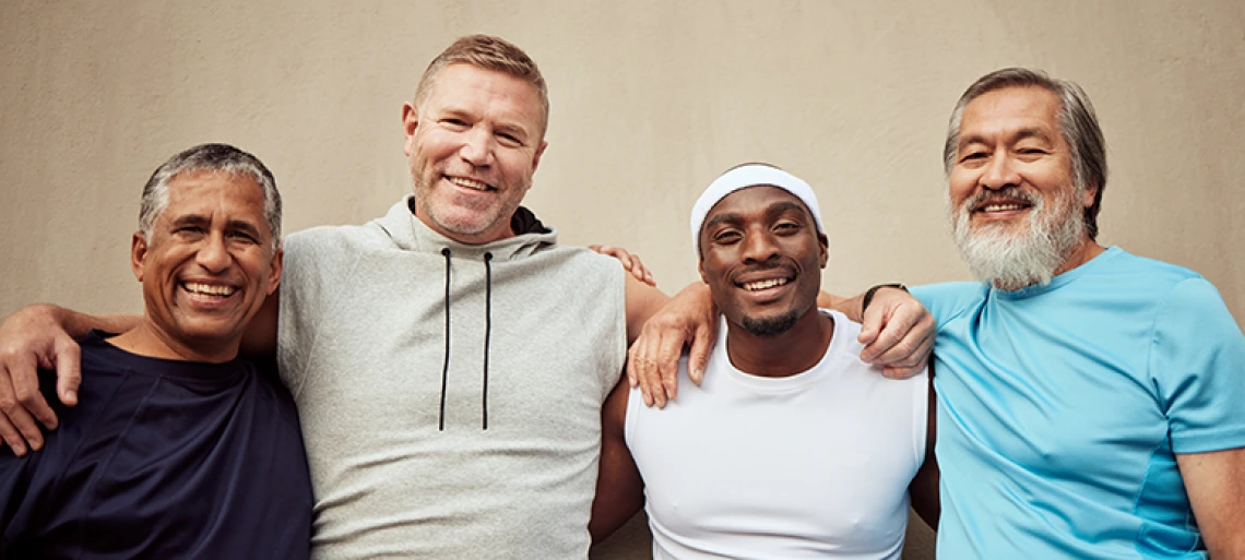Four smiling men in athletic wear stand close together with their arms around each other, posing happily in front of a plain wall.