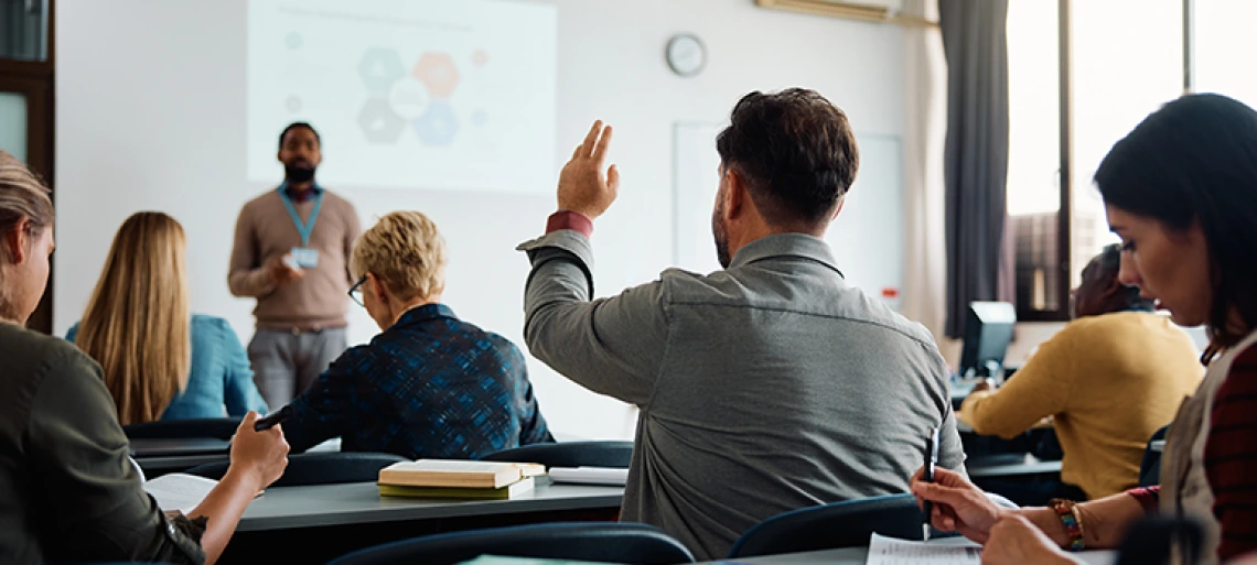 Professional training session with instructor presenting to audience, participant raising hand to ask question in modern classroom setting