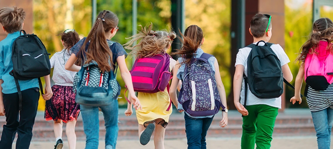 Group of elementary school children with colorful backpacks running together toward school building on a sunny day