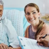 A smiling elderly man in a hospital bed holds hands with a young woman as they talk to a doctor holding a clipboard.